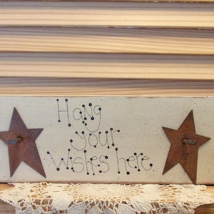 May include: White wooden sign with the text "Hang Your Wishes Here" in black lettering. The sign has two rusty metal stars and four painted hearts in blue, pink, and green.
