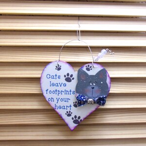 May include: A heart-shaped wooden cat-themed ornament with the text "Cats leave footprints on your heart." The ornament features a painted gray cat with a blue bow tie and paw prints. It hangs from a wire.