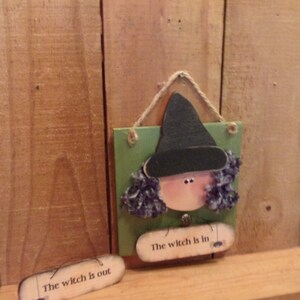 May include: A wooden witch figurine with a black hat and purple hair. The witch is holding a sign that says "The witch is in" with a spider hanging from it. There is a second sign that says "The witch is out" with a spider hanging from it.