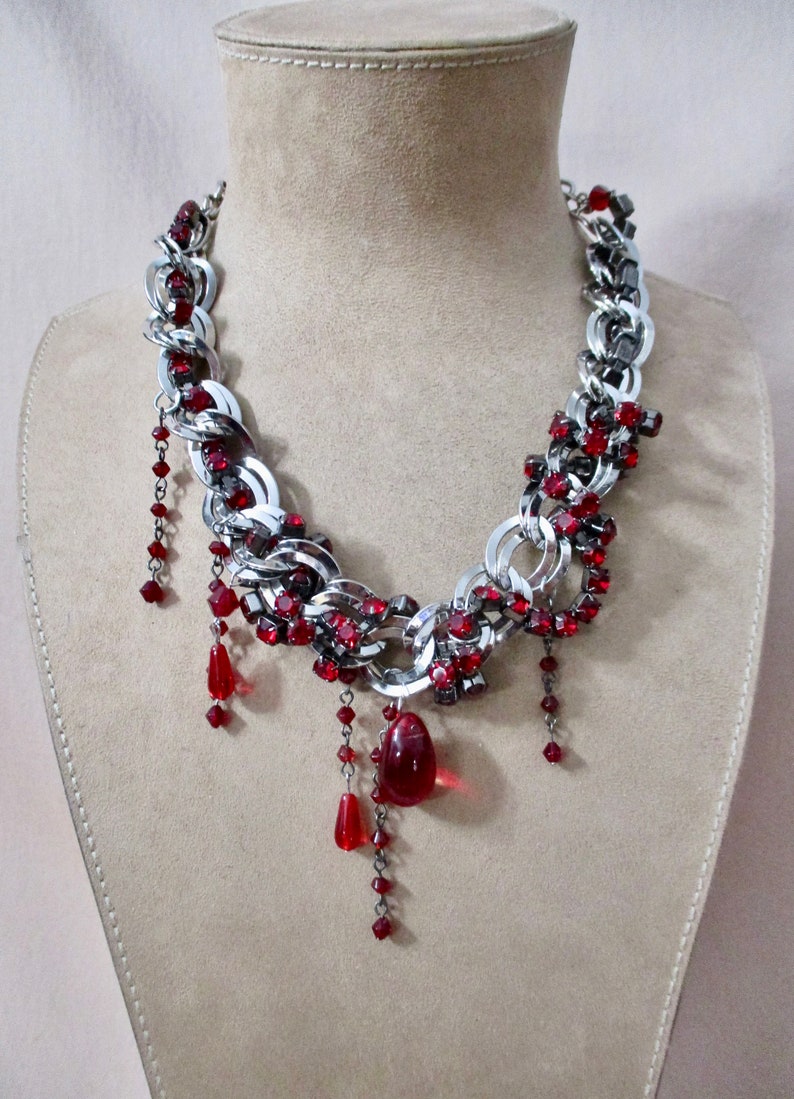 Puede incluir: Un collar de cadena plateada con detalles de cristal rojo y un gran colgante de cristal rojo. El collar est&aacute; en un maniqu&iacute;.