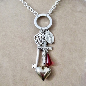 May include: A silver chain necklace with a large silver circle pendant. The pendant has a variety of charms attached to it, including a key, a cross, a heart, and a religious medal. The charms are silver and gold in color.