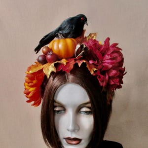 May include: A decorative headpiece featuring a black crow perched atop a small pumpkin and autumn foliage. The headpiece includes red and orange flowers, gold leaves, and small red fruit. The arrangement sits on a mannequin head with brown hair.