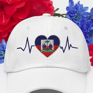 Może przedstawiać: Biała czapka bejsbolowa z haftowanym sercem z flagą Haiti i niebieską linią bicia serca. Czerwone i niebieskie kwiaty w tle.