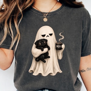Könnte beinhalten: Ein anthrazitfarbenes T-Shirt mit einer Grafik eines beigen Geistes, der einen schwarzen Mops und eine schwarze Kaffeetasse mit der Aufschrift MEH hält. Der Geist hat einen traurigen Gesichtsausdruck und aus der Tasse steigt etwas Dampf auf.