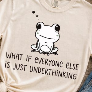 May include: A cream-coloured short-sleeve t-shirt featuring a black and white line drawing of a sitting frog with large eyes and the text "WHAT IF EVERYONE ELSE IS JUST UNDERTHINKING" printed in black lettering below the graphic illustration.