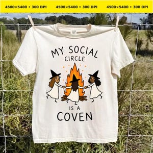 May include: A cream-colored t-shirt featuring a graphic of three figures in black witch hats and white dresses holding hands around a campfire. The text above the graphic reads "MY SOCIAL CIRCLE" and the text below reads "IS A COVEN" in black.