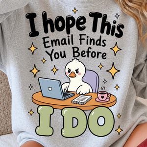 Op de afbeelding: Een lichtgrijs sweatshirt met een afbeelding van een witte eend die aan een oranje bureau zit met een laptop, een notitieblok en een roze koffiekopje. De tekst luidt I hope This Email Finds You Before I DO, omringd door kleine gele sterren.