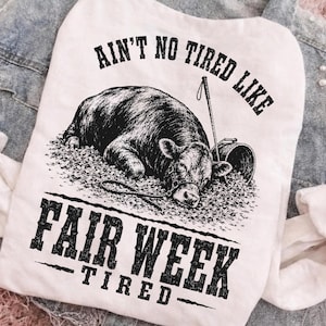 Può includere: Una t-shirt bianca con una grafica nera di una mucca sdraiata sulla lettiera, insieme a un bastone da esposizione e un secchio. Il testo AIN'T NO TIRED LIKE FAIR WEEK TIRED è stampato in un carattere nero usurato sul davanti del capo.