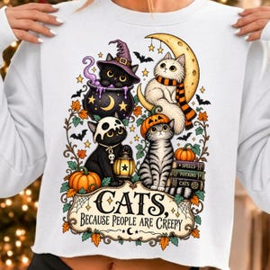 May include: A white sweatshirt featuring a graphic of four cats in Halloween attire, including a black cat in a witch hat, a white cat on a crescent moon, and two cats with pumpkins. The text reads, "CATS, BECAUSE PEOPLE ARE CREEPY."