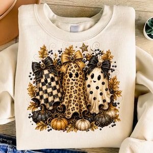 May include: A cream-colored crewneck sweatshirt featuring a graphic of three ghosts wearing black bows. The ghosts have checkered, leopard print, and polka dot patterns. They are surrounded by autumn leaves, berries, and small pumpkins.
