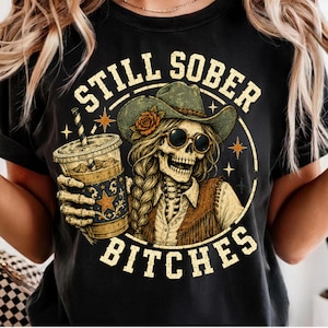 May include: A black t-shirt featuring a graphic of a skeleton wearing a cowboy hat with a rose, sunglasses, and a fringed vest. The skeleton holds a drink with a straw. The text STILL SOBER BITCHES is printed in a circular arrangement around the design.