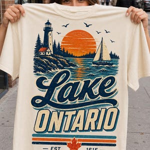 May include: A cream-colored t-shirt featuring a graphic of a lighthouse, a sailboat, and a large orange sun over water. The text reads Lake Ontario, Est. 1615, with a red maple leaf icon. The design uses blue, orange, and cream colors throughout.