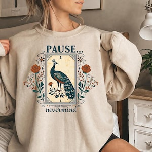 May include: A beige crewneck sweatshirt featuring a graphic print of a peacock inside a rectangular frame. The text reads PAUSE... nevermind. The design includes floral elements in orange and green, surrounding the central peacock illustration.