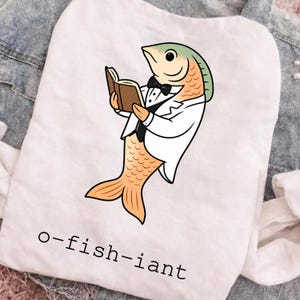 以下が含まれることがあります： 白いTシャツに、白いタキシードジャケット、黒いベスト、黒い蝶ネクタイを身につけ、茶色の本を読んでいるオレンジ色の魚のグラフィックが描かれています。魚の下には、シンプルな黒のサンセリフフォントで「o-fish-iant」という文字がプリントされています。