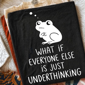 May include: A black t-shirt featuring a white graphic of a frog with a thought bubble above its head. Below the frog, the text reads: WHAT IF EVERYONE ELSE IS JUST UNDERTHINKING. The shirt is folded and layered over other garments.