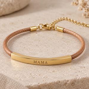 Puede incluir: Una pulsera de cordón de cuero color canela con una barra metálica dorada curvada grabada con la palabra MAMA. La pulsera incluye una cadena extensora dorada con un cierre de mosquetón y una pequeña etiqueta metálica al final de la cadena.