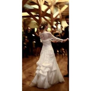 May include: A floor-length white wedding dress featuring long lace sleeves, a fitted bodice, and a voluminous skirt with layered, ruffled fabric detailing. The gown has a structured silhouette that flares toward the hem, creating a full shape.