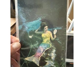 May include: A vintage postcard showing a mermaid with dark hair, wearing a yellow top and a shimmering, multicoloured tail. She holds a mirror and is surrounded by underwater elements, with a dark background and blue water effects.