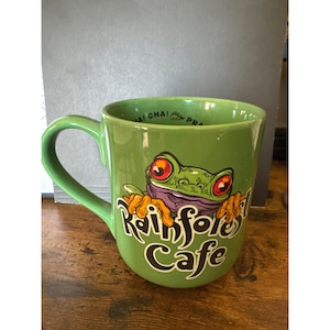 May include: Green ceramic mug with a large handle. The mug features a cartoon frog with red eyes and the words "Rainforest Cafe" in a stylized font. The mug is sitting on a wooden surface.