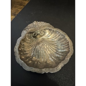 May include: A silver-toned, shell-shaped dish with a scalloped edge. The dish has a detailed, textured surface with a central spiral design. The top edge features a decorative floral pattern. This vintage piece is ideal for serving or display.