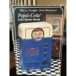 May include: A Pepsi-Cola coin sorter bank in its original box. The bank is blue with the Pepsi-Cola logo and text. The box has a black and white checkered pattern on the bottom.