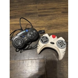 May include: Two vintage video game controllers. One is black with a D-pad, buttons, and a blue button. The other is white with gray and red accents, featuring a unique design. Both controllers have attached cords.