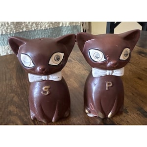 May include: A pair of vintage brown ceramic cat-shaped salt and pepper shakers. Each cat has large, wide eyes, a white bow tie, and a letter on its chest. One shaker has an "S" and the other has a "P".
