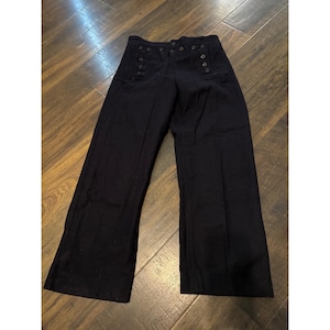 May include: A pair of dark navy blue high-waisted trousers featuring a wide-leg silhouette and a distinctive double-breasted button front closure. The pants have a structured waistband with two vertical rows of dark buttons, creating a classic sailor-inspired aesthetic with clean lines and a solid color finish.