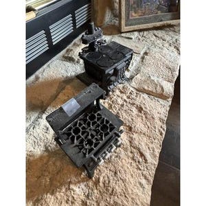 Puede incluir: Dos miniaturas de estufas de hierro fundido negro. Una estufa tiene cuatro quemadores circulares y una chimenea. La otra estufa tiene una placa de cocina de seis quemadores. Ambas estufas están sobre una superficie de piedra.