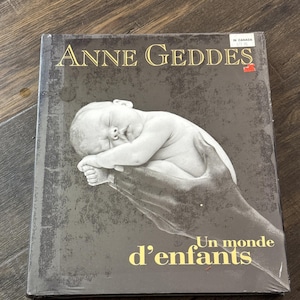 May include: A book titled "Anne Geddes" with a black and white image of a sleeping baby cradled in a hand. The book cover has gold lettering and the French title "Un monde d'enfants".