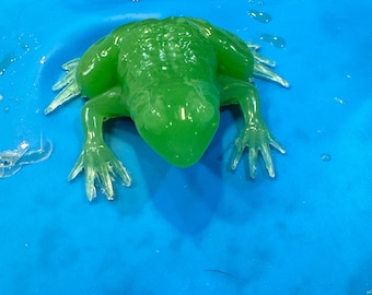 Puede incluir: Una figurita de rana verde brillante hecha de resina transparente. La rana está sentada sobre una superficie azul.