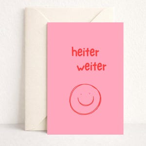Könnte beinhalten: Eine rosa Grußkarte mit den Worten "heiter weiter" in Rot und einem roten Smiley. Dazu gehört ein cremefarbener Umschlag. Das Design ist einfach und fröhlich, passend für verschiedene Anlässe.