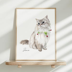 K&ouml;nnte beinhalten: Ein gerahmter Aquarelldruck eines flauschigen, hellen Katzenportr&auml;ts mit grauen Abzeichen, das ein gr&uuml;n-wei&szlig;es Halsband mit kleinen roten Akzenten tr&auml;gt. Der Name Juno steht in schwarzer Schreibschrift unter der Katze auf wei&szlig;em Grund.