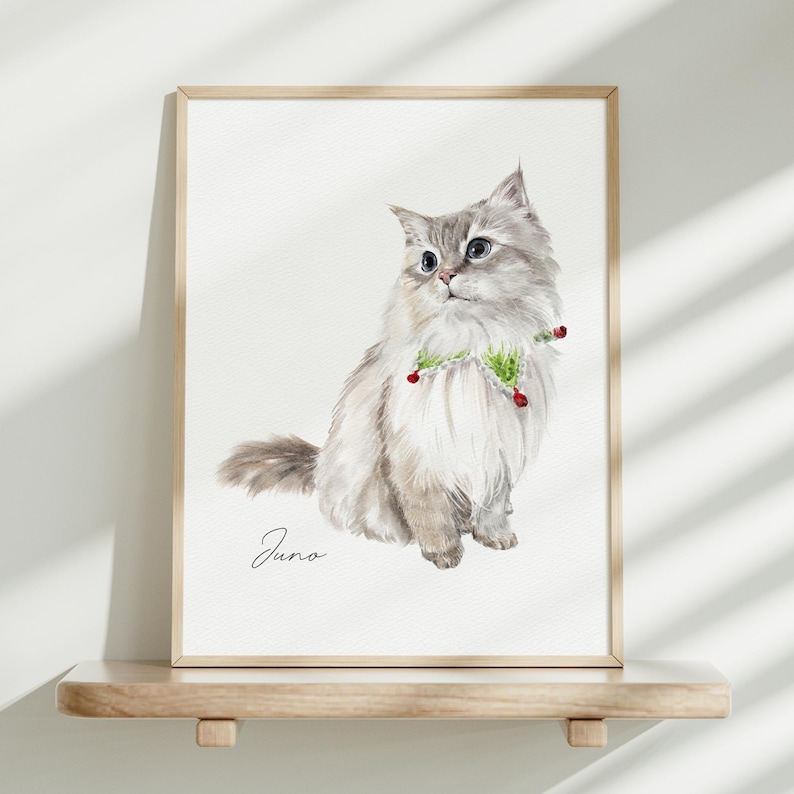 K&ouml;nnte beinhalten: Ein gerahmter Aquarelldruck eines flauschigen, hellen Katzenportr&auml;ts mit grauen Abzeichen, das ein gr&uuml;n-wei&szlig;es Halsband mit kleinen roten Akzenten tr&auml;gt. Der Name Juno steht in schwarzer Schreibschrift unter der Katze auf wei&szlig;em Grund.