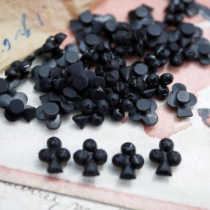 May include: A collection of black plastic buttons shaped like playing card clubs. The buttons are scattered on a white surface with a brown paper background.