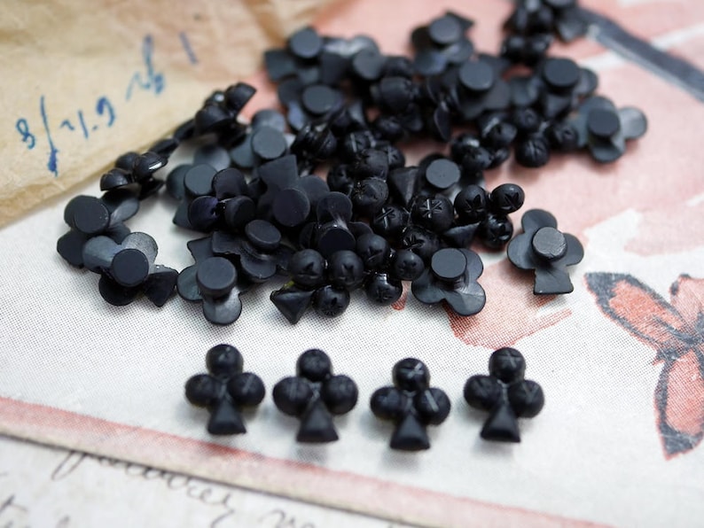 May include: A collection of black plastic buttons shaped like playing card clubs. The buttons are scattered on a white surface with a brown paper background.