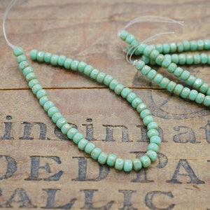 May include: A strand of light blue glass beads with a faceted finish. The beads are strung on a clear thread and are arranged in a circular pattern. The beads are displayed on a wooden surface.