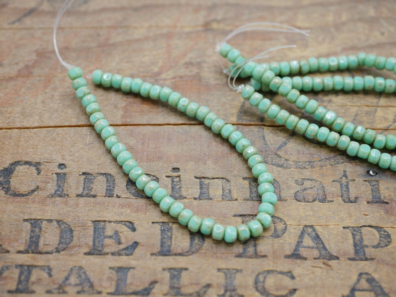 May include: A strand of light blue glass beads with a faceted finish. The beads are strung on a clear thread and are arranged in a circular pattern. The beads are displayed on a wooden surface.
