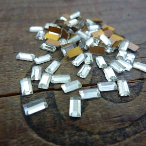 May include: A pile of clear glass rectangular rhinestones on a wooden surface.