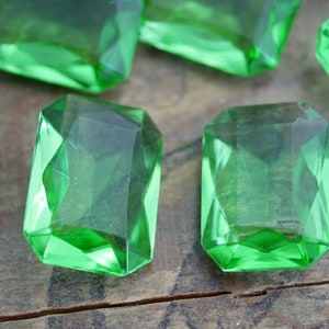 May include: Six emerald green glass gems with a faceted cut. The gems are arranged on a wooden surface.