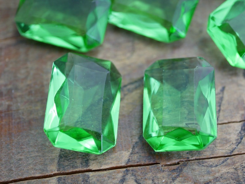 May include: Six emerald green glass gems with a faceted cut. The gems are arranged on a wooden surface.