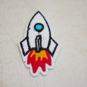 May include: Embroidered patch of a white rocket ship with black outlines. The rocket has a blue porthole and red and yellow flames. This patch is ideal for decorating clothing or accessories.