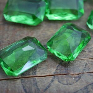 May include: A group of emerald green glass gems, cut into rectangular shapes, are scattered on a wooden surface.