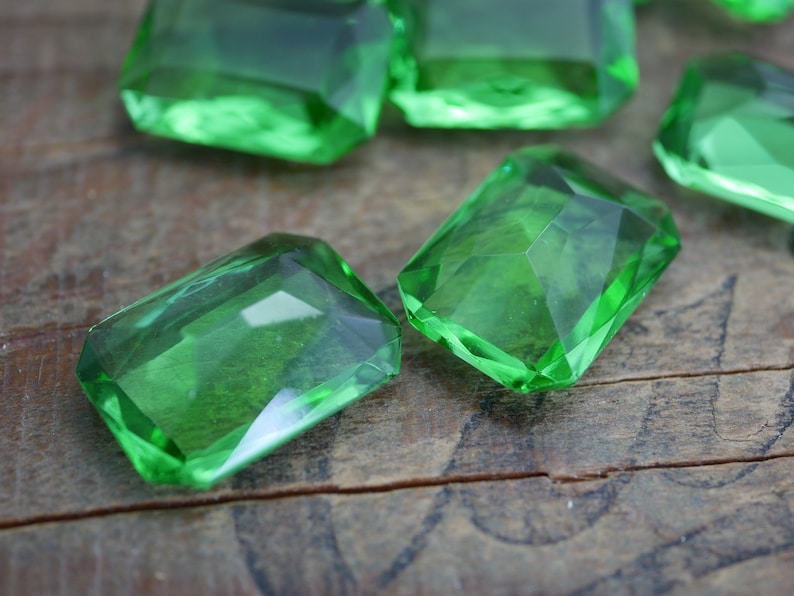May include: A group of emerald green glass gems, cut into rectangular shapes, are scattered on a wooden surface.
