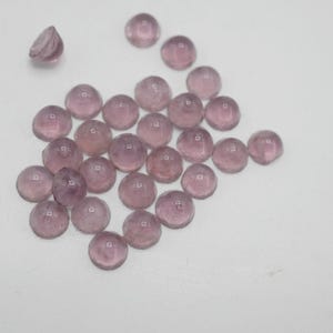 Op de afbeelding: Een verzameling ronde, doorschijnende, lichtpaarse glazen cabochons. De cabochons zijn verspreid over een wit oppervlak.