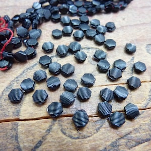 以下が含まれることがあります： テクスチャ加工された表面を持つ黒い六角形のガラスビーズのクローズアップ。ビーズは木製の表面に散らばっています。