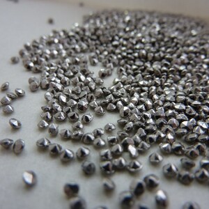 May include: A close-up image shows a large quantity of small, silver-coloured, faceted beads. The beads are spread across a light surface, reflecting light and creating a metallic shine. These beads are likely used for jewellery making or craft projects.