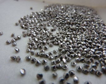 May include: A close-up shot displays a multitude of small, silver-colored, faceted beads. The beads are scattered across a light-colored surface, catching the light and reflecting a metallic sheen. The beads are likely used for jewelry making or craft projects.