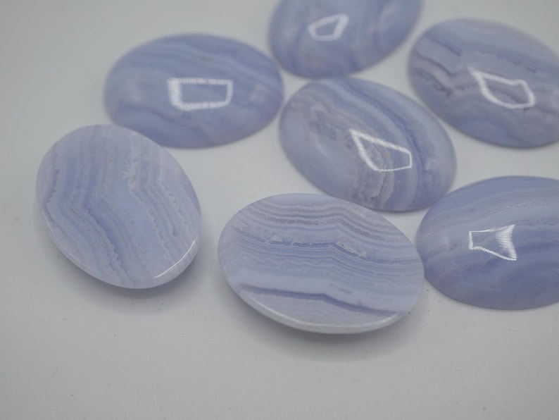 May include: A collection of oval-shaped, pale blue gemstones with swirling patterns. The polished stones have a smooth, glossy surface and vary slightly in their color and veining. These stones are likely used for jewelry or crafts.