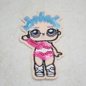 May include: Embroidered patch of a cartoon doll with blue sequin hair, large purple eyes, and a pink sequin top. The doll wears white trainers with star details. The patch is on a beige background.